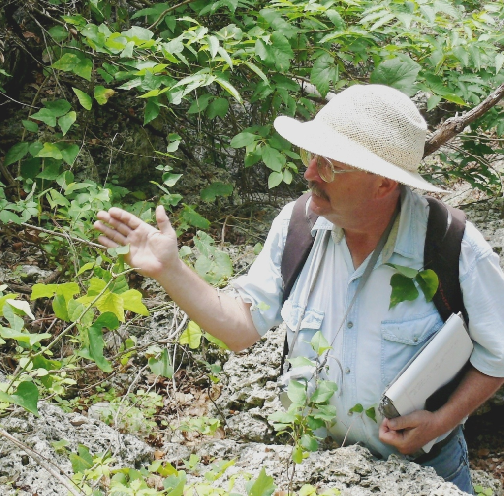 A person in a hat and glasses, doing field work, observing a plant and taking notes