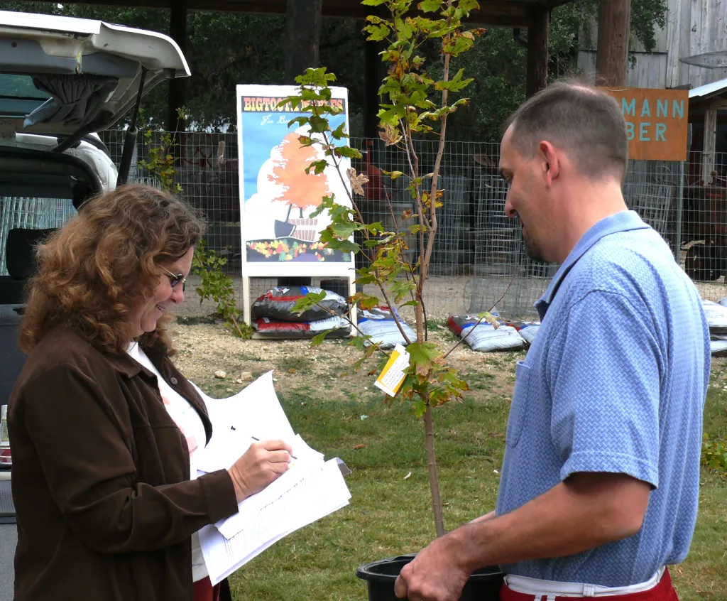 Two people at an event outside; one of them is hoding a potted plant and the other is marking a piece of paper.